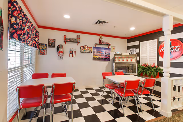 A retro-style dining area with black and white checkered floor tiles, red chairs, and white tables. The walls are decorated with vintage signs and memorabilia, including a Coca-Cola sign and Route 66 themed items. Large windows with blinds and patterned valances let in natural light.