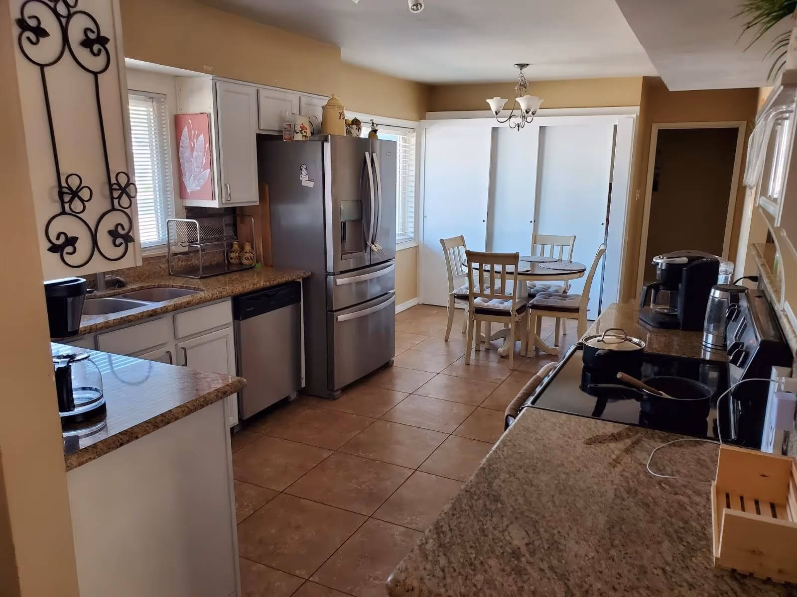 A bright kitchen with beige walls and tiled floor featuring granite countertops, white cabinets, a stainless steel refrigerator, dishwasher, and stove. A small round dining table with four chairs is placed near a window with white blinds and sliding closet doors. Various kitchen appliances and utensils are visible on the counters.