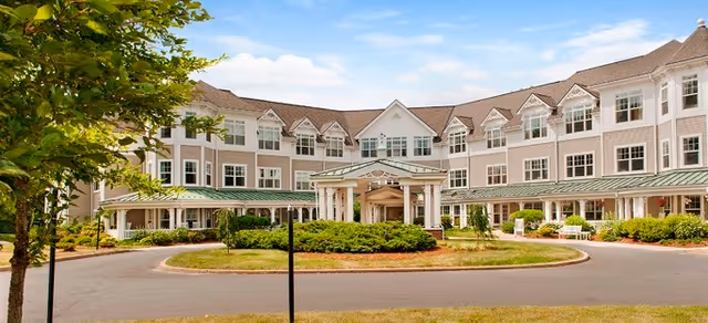Front exterior view of a large, three-story senior living facility building with multiple windows, a covered entrance with white columns, surrounded by greenery and a circular driveway under a partly cloudy sky.