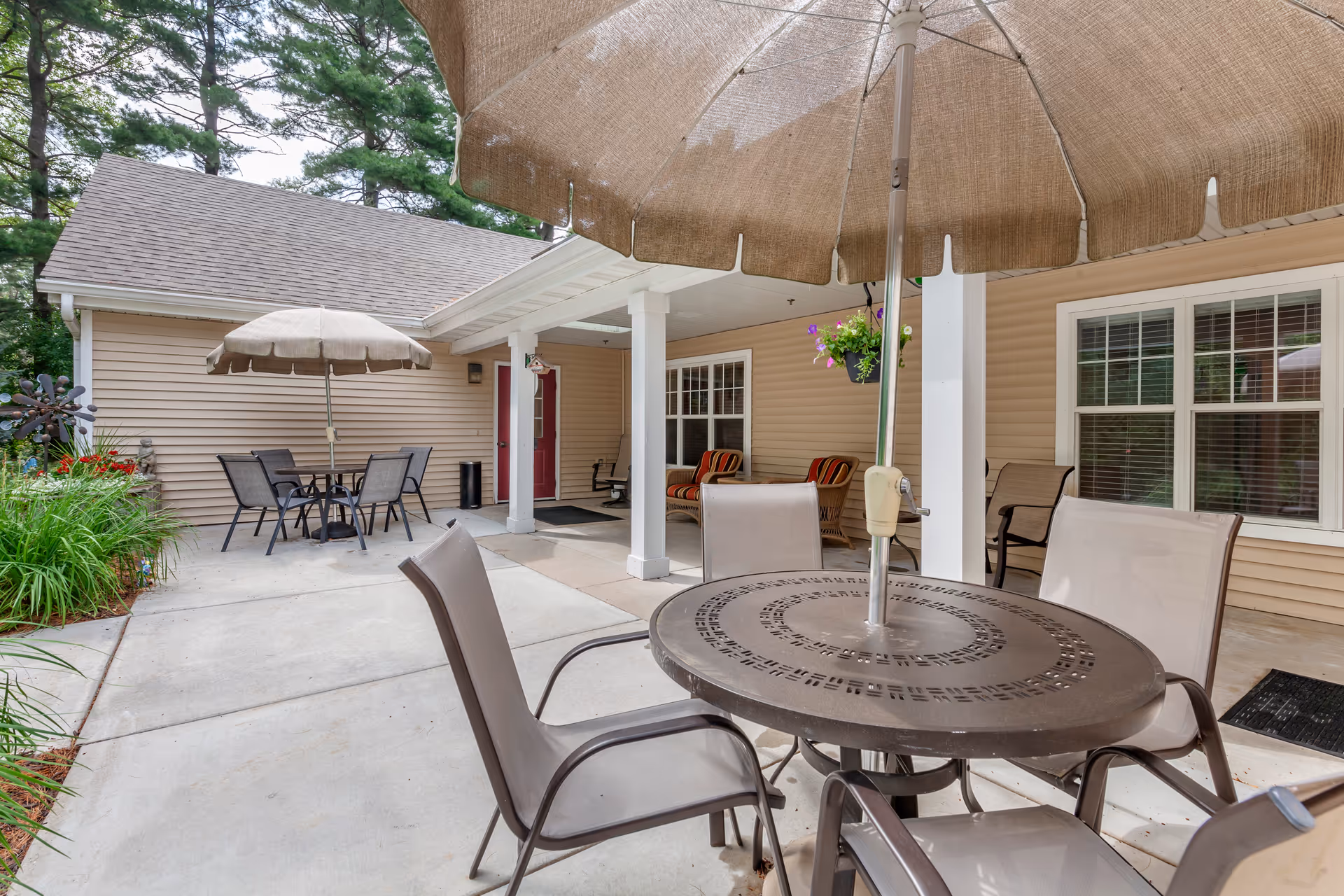 Outdoor patio area with round metal tables, umbrellas and chairs in front of a beige siding building with a covered porch.