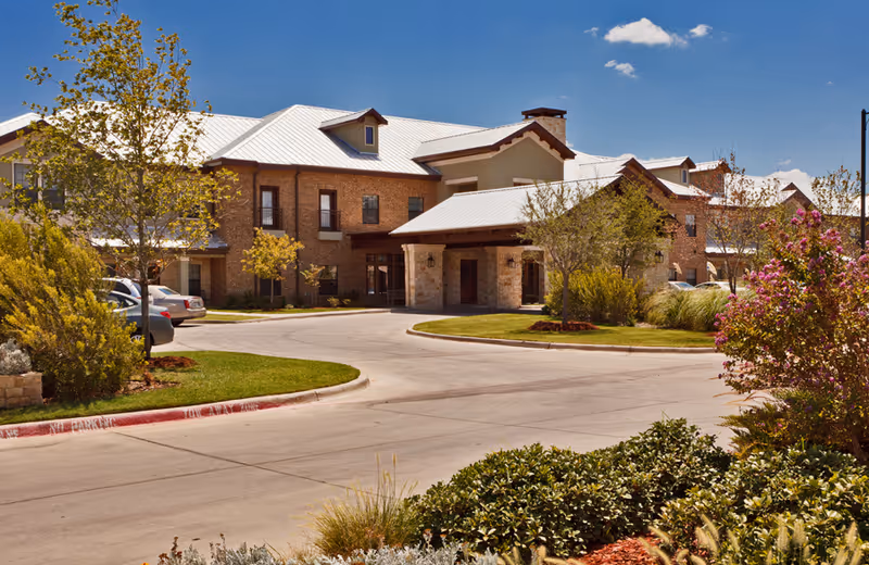 Exterior view of a senior living facility named Waterview The Point Independent Living, showing a large brick building with a metal roof, surrounded by landscaped greenery, trees, and a driveway with parked cars under a clear blue sky.