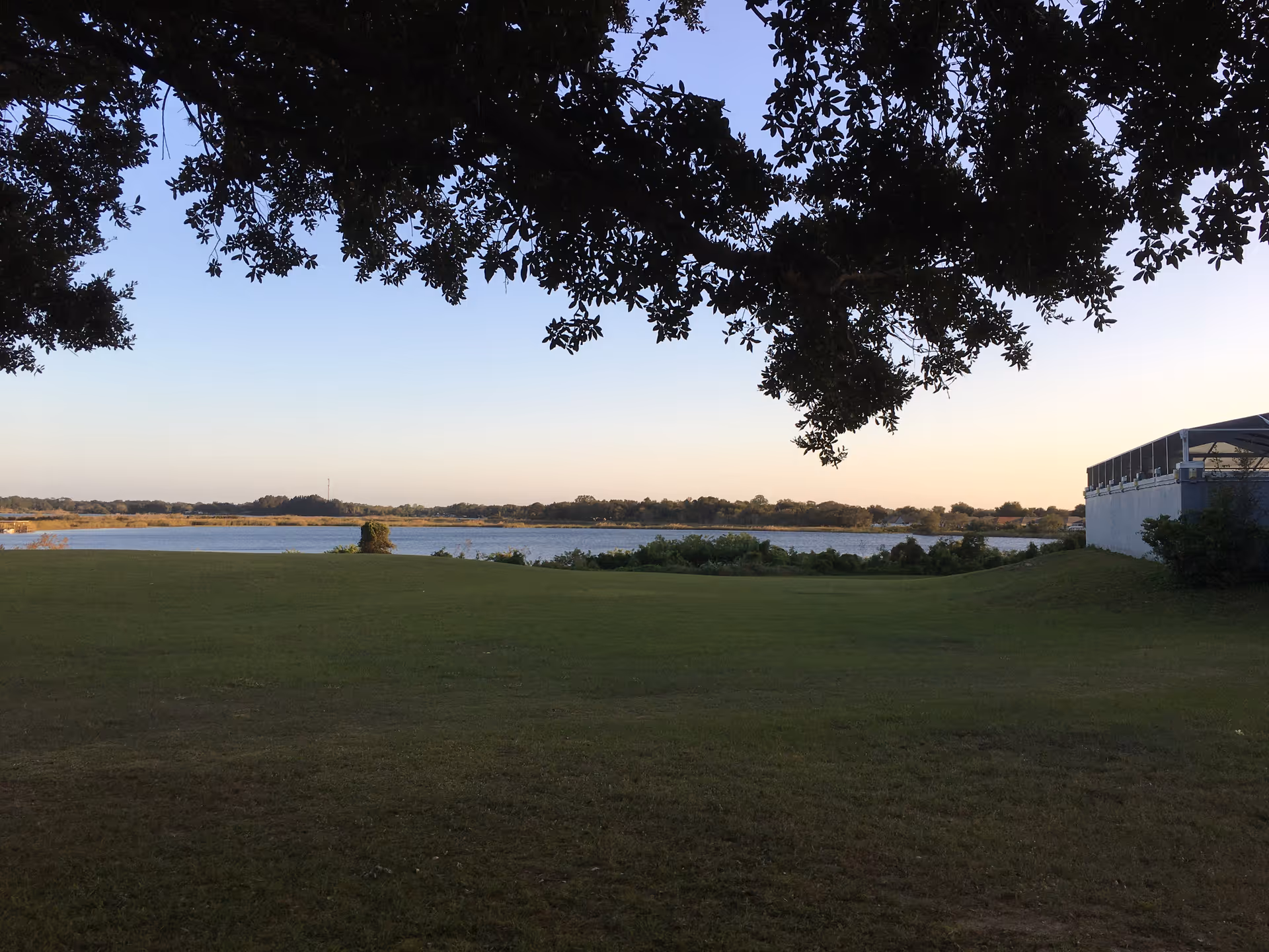 A scenic outdoor view featuring a grassy lawn leading to a body of water with trees and shrubs along the shoreline. The sky is clear with a soft gradient from light blue to pale orange, indicating either sunrise or sunset. Tree branches with leaves frame the top of the image, and a building with a covered patio is visible on the right side.
