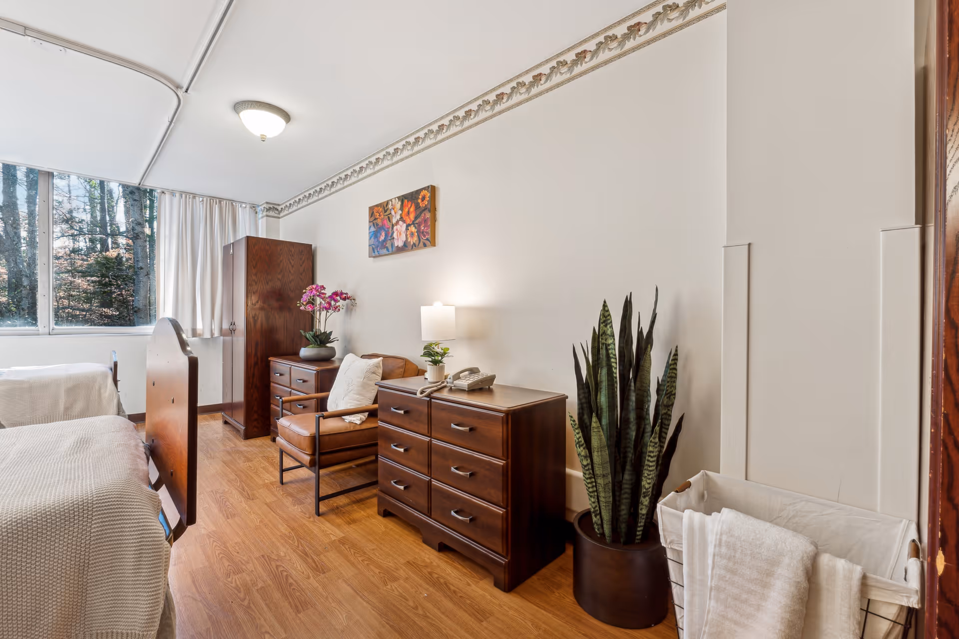 Sunny nursing facility bedroom with two beds, wooden dressers, an armchair, potted plants and a window overlooking trees.