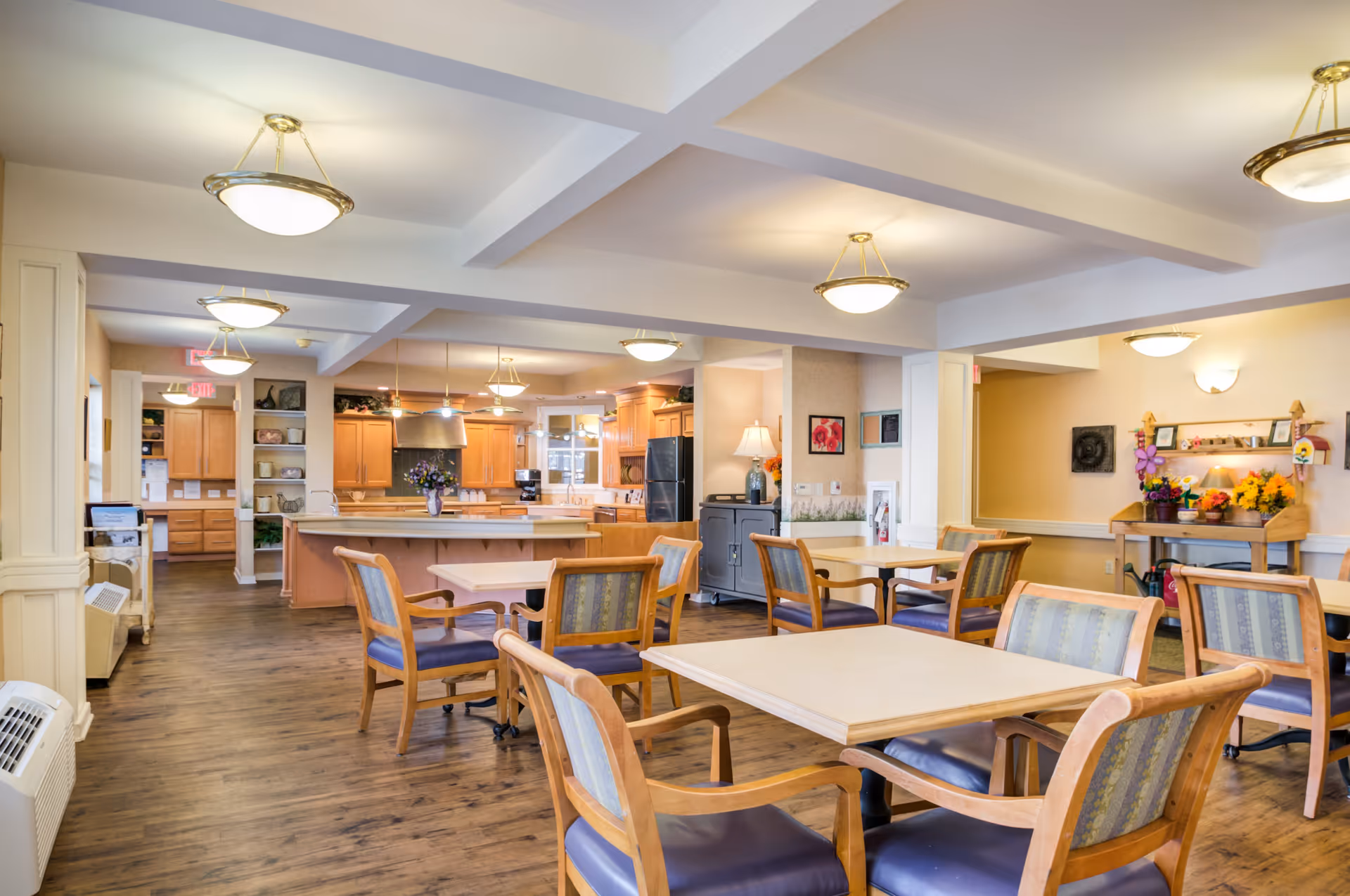 A bright and spacious dining area in a senior living facility featuring multiple wooden tables with cushioned chairs arranged around them. The room has warm lighting from ceiling fixtures and a wooden floor. In the background, there is a large open kitchen with wooden cabinets, a kitchen island, and modern appliances. The walls are decorated with framed artwork and a small table with flowers and decorative items.