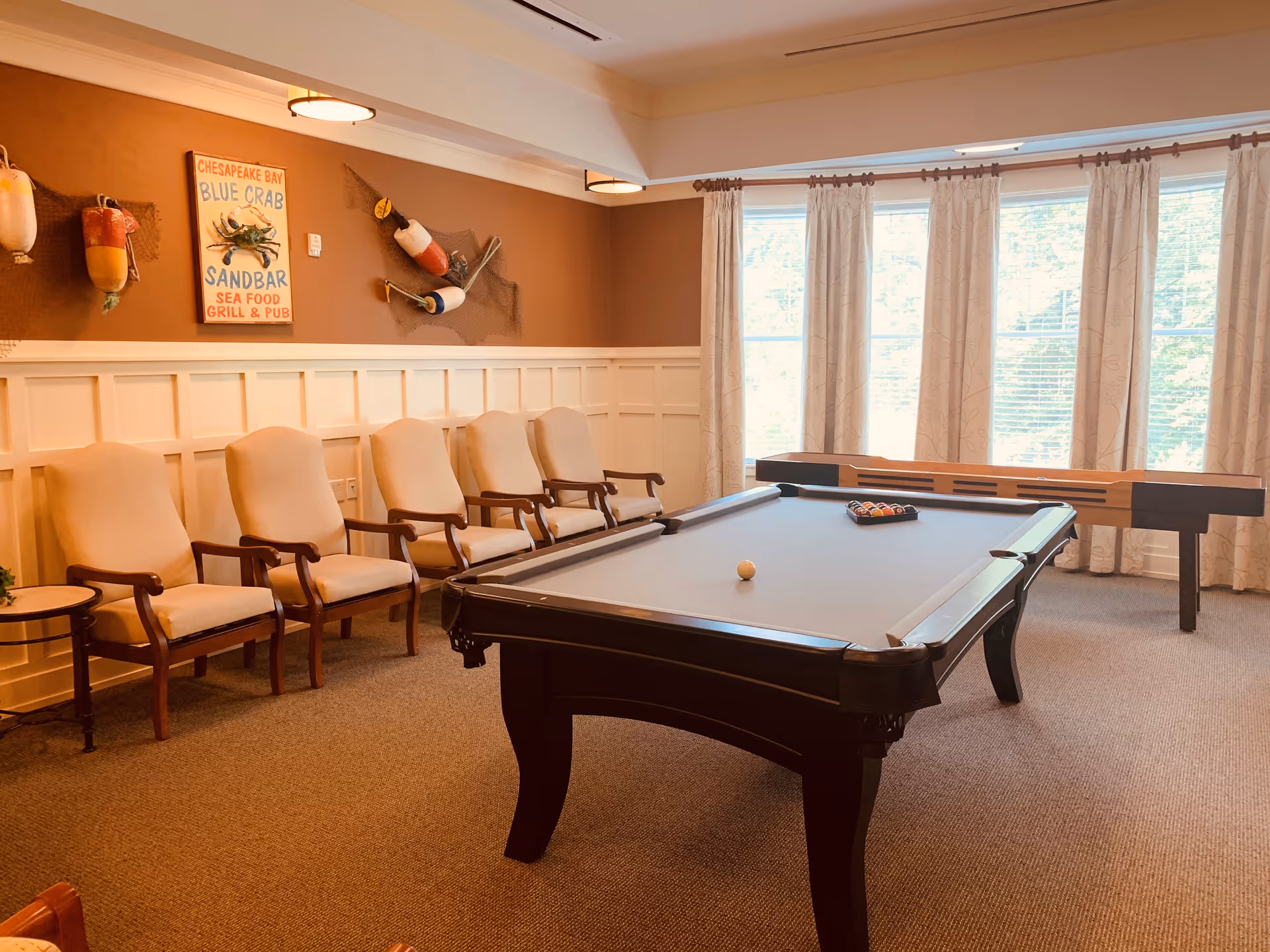 A recreational room with a pool table and a shuffleboard table near large windows with curtains. There are five beige cushioned chairs with wooden armrests lined up against a wall decorated with fishing buoys and a Chesapeake Bay Blue Crab seafood grill and pub sign.