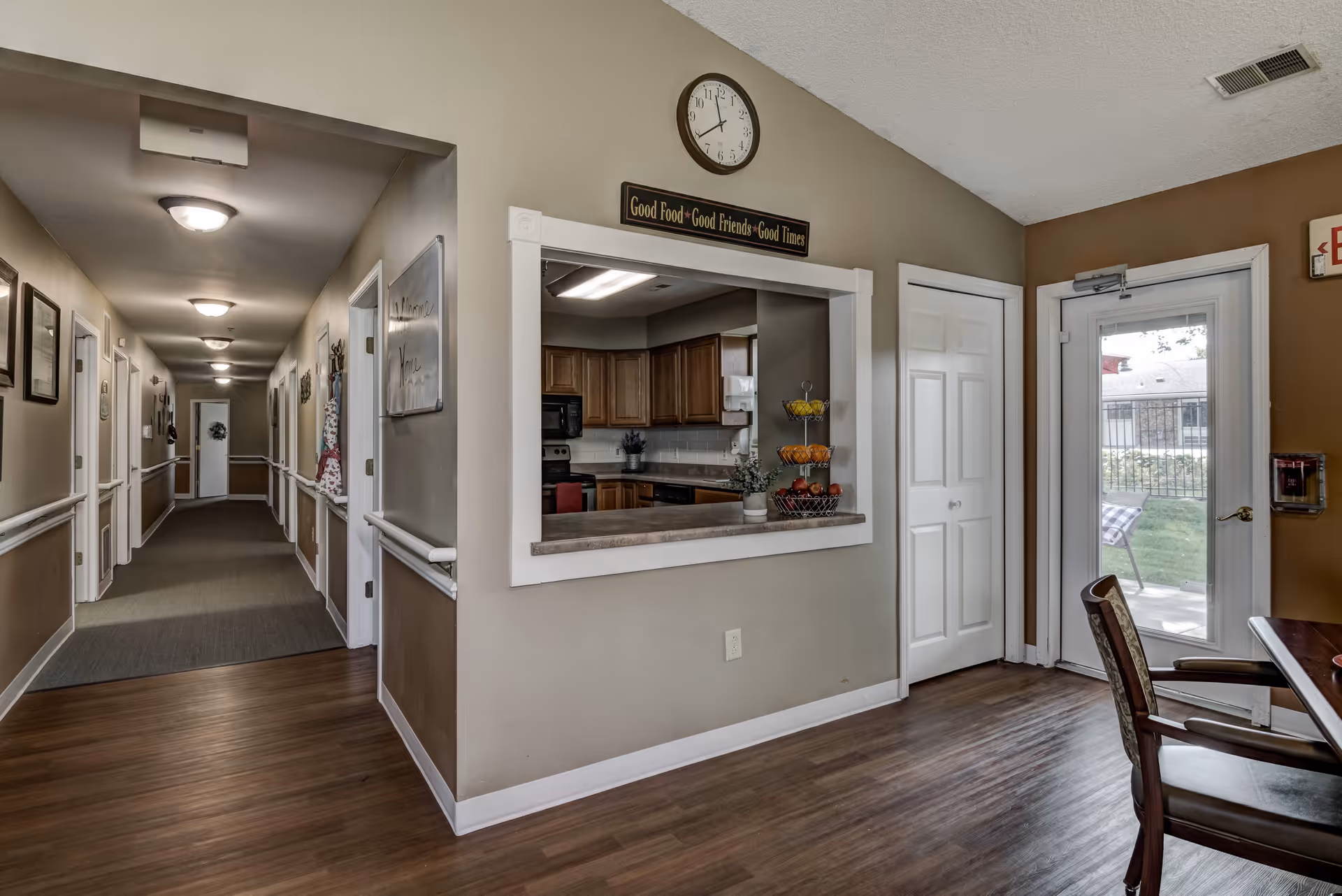 Interior view of a senior living facility hallway with beige walls and wooden flooring. To the right, there is a kitchen area visible through a serving window with wooden cabinets and a countertop holding a tiered fruit basket. Above the window is a clock and a sign that reads 'Good Food • Good Friends • Good Times'. A door with a glass window leads outside to a patio area with a bench and greenery. A chair and part of a table are visible in the foreground.