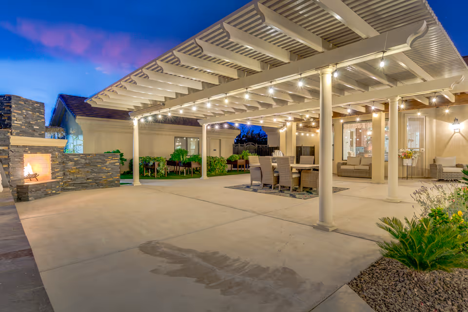 Outdoor patio area at dusk with a pergola overhead adorned with string lights. There is a stone fireplace on the left side with a fire burning. Under the pergola, there is a dining table with six wicker chairs on a rug, and in the background, there are cushioned seating areas near the building. Some plants and landscaping are visible around the patio.