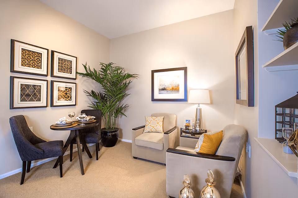 Cozy living room area with two armchairs, a small round dining table with two chairs, framed wall art, a floor plant, and decorative accents.