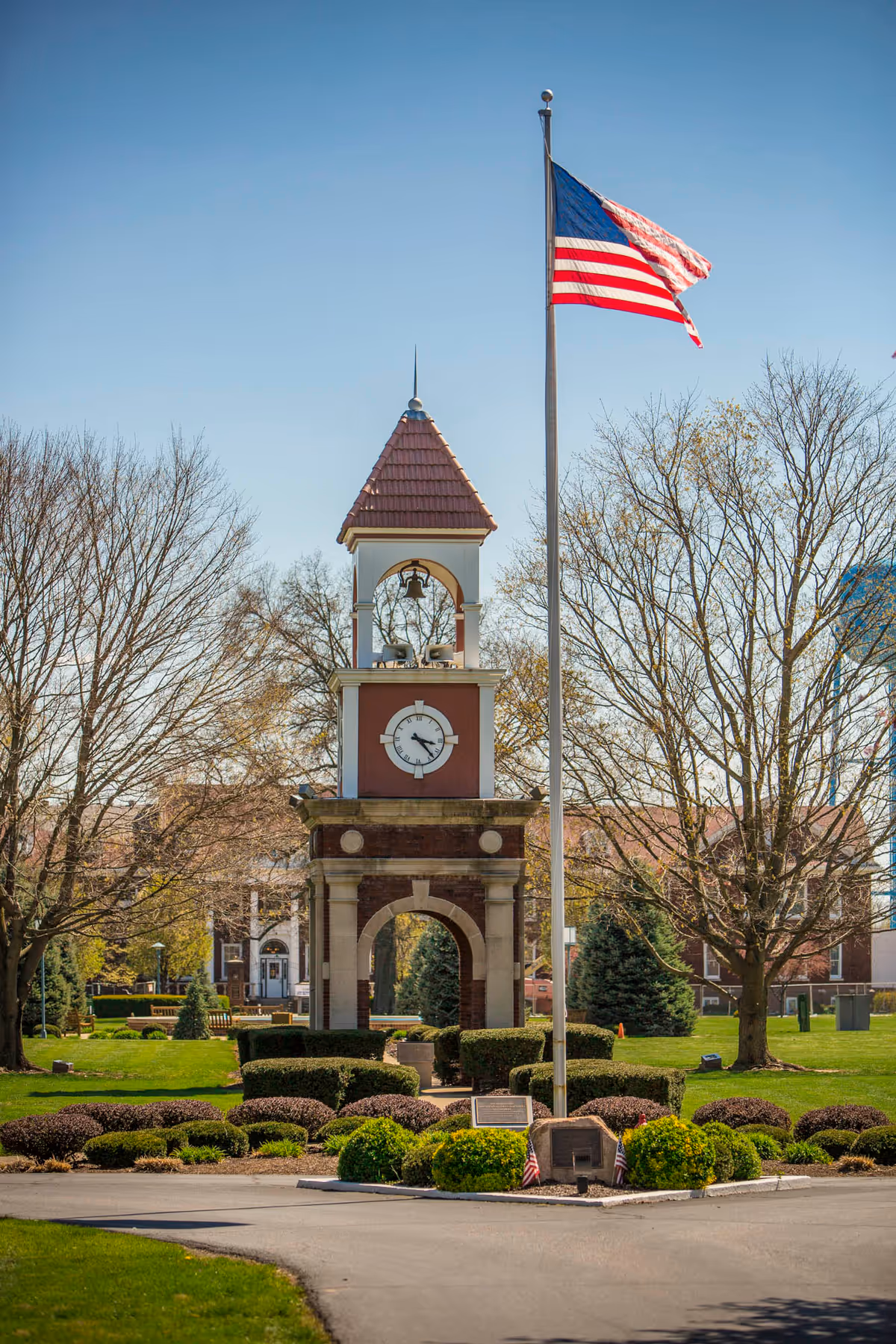 A clock tower with a peaked roof and bell, surrounded by neatly trimmed bushes and trees with sparse leaves. An American flag on a tall flagpole is prominently displayed in front of the tower. The scene is set in a well-maintained outdoor area with a clear blue sky.