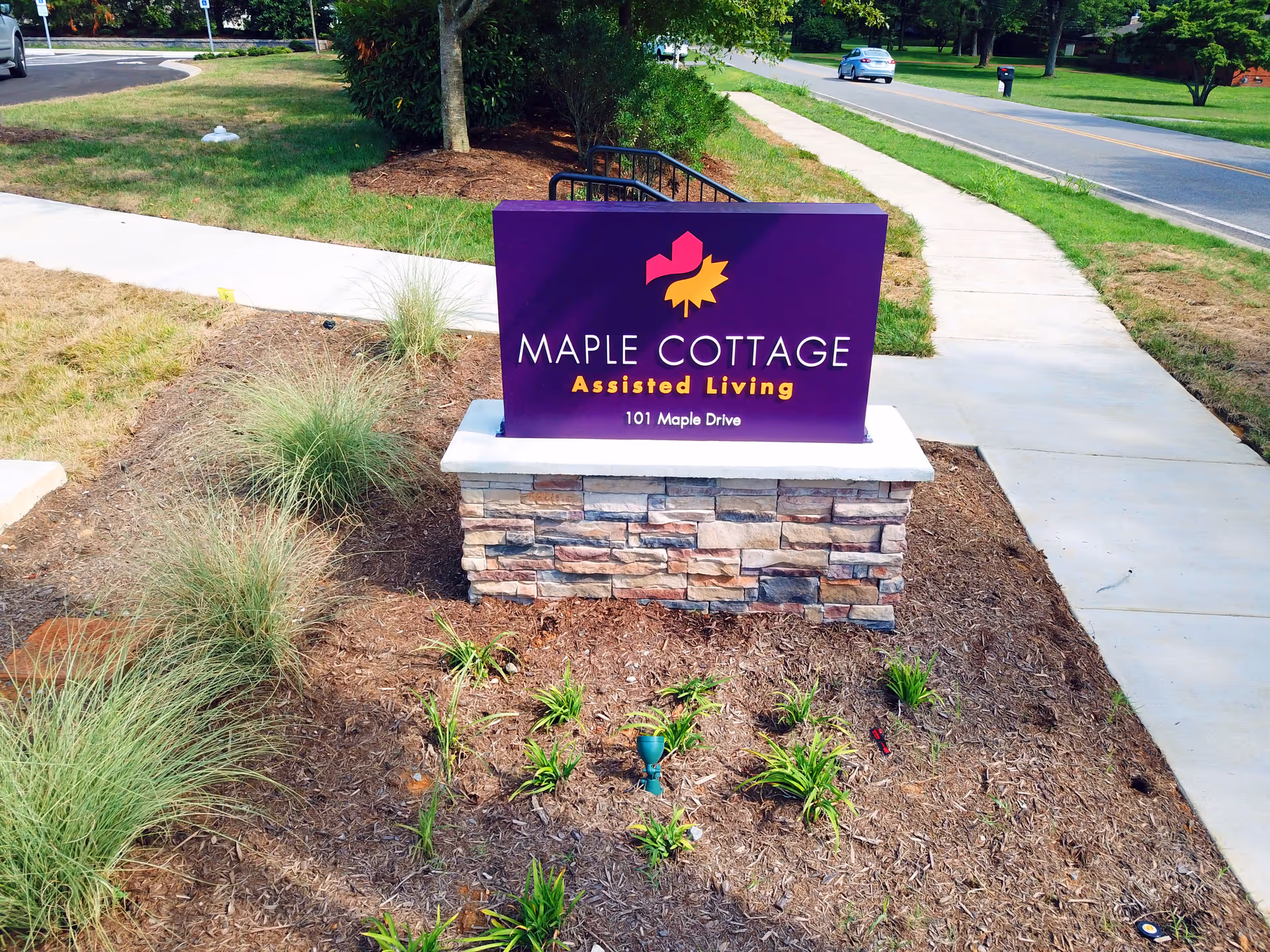 Outdoor view of a purple sign for Maple Cottage Assisted Living at 101 Maple Drive, mounted on a stone base surrounded by landscaping with grass and small plants, next to a sidewalk and a road with a car in the background.