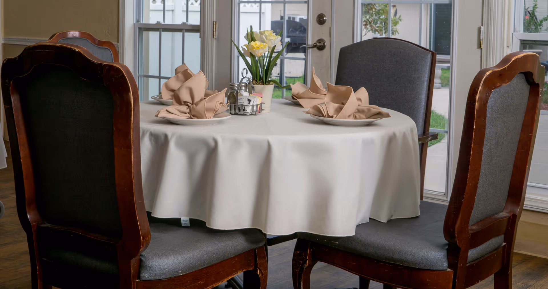 A round dining table covered with a white tablecloth set with four plates, each with a folded beige napkin. In the center of the table is a small flower arrangement with yellow and white flowers, along with salt and pepper shakers and a condiment holder. The table is surrounded by four wooden chairs with gray cushions. Behind the table are large windows and a glass door leading outside.