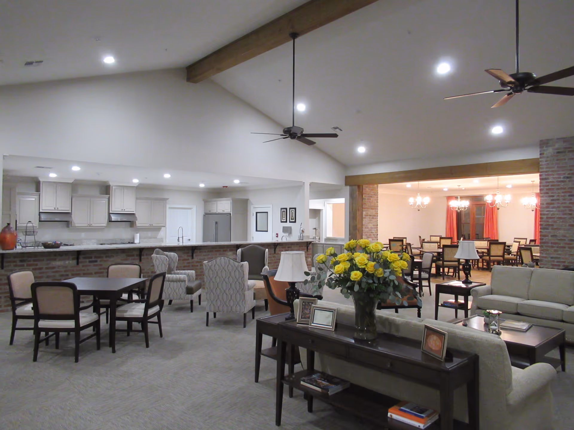 Spacious senior living common area featuring a kitchen with white cabinets and stainless steel appliances, multiple seating arrangements including armchairs and dining tables, a sofa set with a coffee table, and a large bouquet of yellow roses on a console table. The room has high vaulted ceilings with exposed wooden beams and ceiling fans, and a dining room with chandeliers and red curtains is visible in the background.
