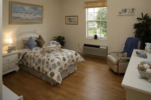 Neat, well-lit single bedroom with a bed, nightstand, armchair, dresser, and window overlooking greenery.