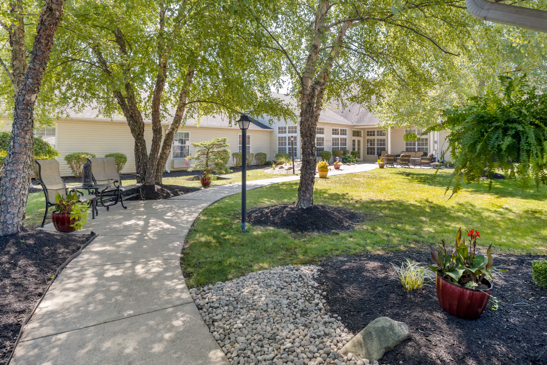 A sunny outdoor garden area at Brookdale Piqua featuring a curved concrete walkway, several trees with string lights, benches, potted plants, and a building with large windows in the background.