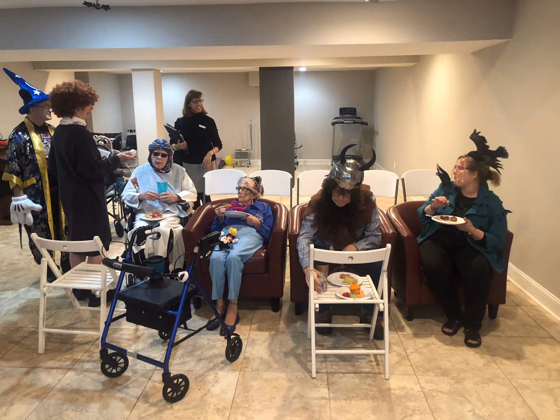 A group of elderly people and caregivers in a room, some seated in armchairs and others standing. Several individuals are wearing festive costumes and hats, including a wizard hat, a Viking helmet, and a witch hat. Some people are holding plates of food, and there is a walker in front of one seated person. The room has tiled floors and neutral-colored walls with a treadmill in the background.