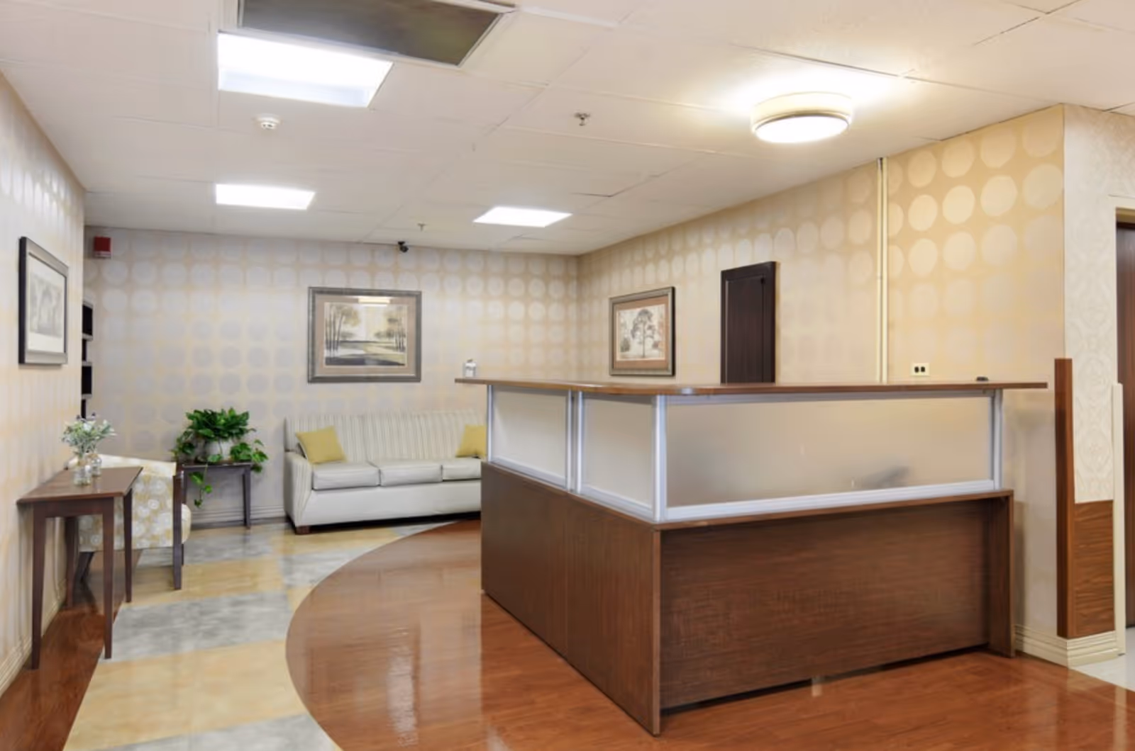 Reception area in BRIA of Chicago Heights featuring a wooden reception desk with frosted glass panels, a beige sofa with yellow cushions, framed artwork on the walls, a small table with a plant, and patterned wallpaper with circular designs.