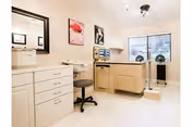 Bright clinical-style room with beige cabinets and counters, a sink and mirror, a rolling stool, medical equipment, and a large window.