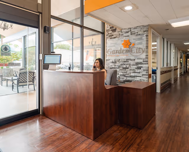 Reception area of Garden City Healthcare Center with a woman sitting behind a wooden front desk, talking on the phone. Behind her is a stone accent wall with the Garden City Healthcare Center logo. To the left, there is a glass door leading to an outdoor seating area with chairs and tables. A hallway extends to the right.