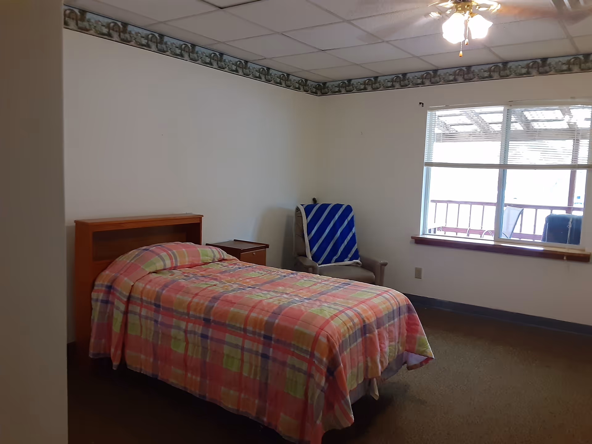 A simple bedroom with a single bed covered in a colorful plaid blanket, a wooden headboard, a small wooden nightstand, a beige armchair with a blue and white striped blanket draped over it, a large window with blinds, and a ceiling fan with lights. The walls are white with a decorative border near the ceiling.