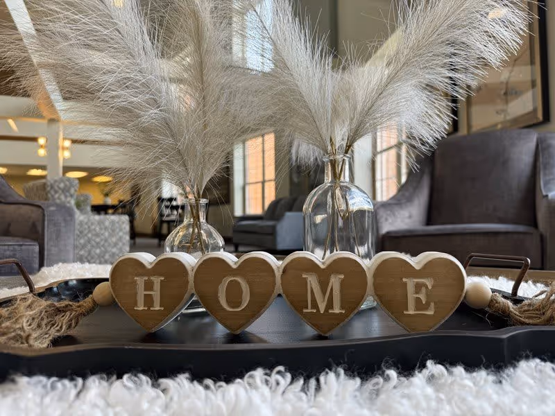 Close-up of a decorative tray on a table with wooden heart-shaped blocks spelling the word HOME, two glass vases with pampas grass, and a cozy living room with armchairs and large windows in the background.
