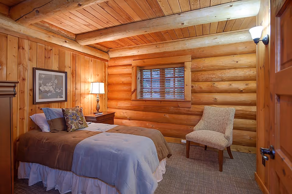 Cozy bedroom with log cabin style wooden walls and ceiling beams, featuring a bed with brown and light blue bedding, a nightstand with a lamp, a framed picture on the wall, a window with wooden blinds, and a beige upholstered chair.