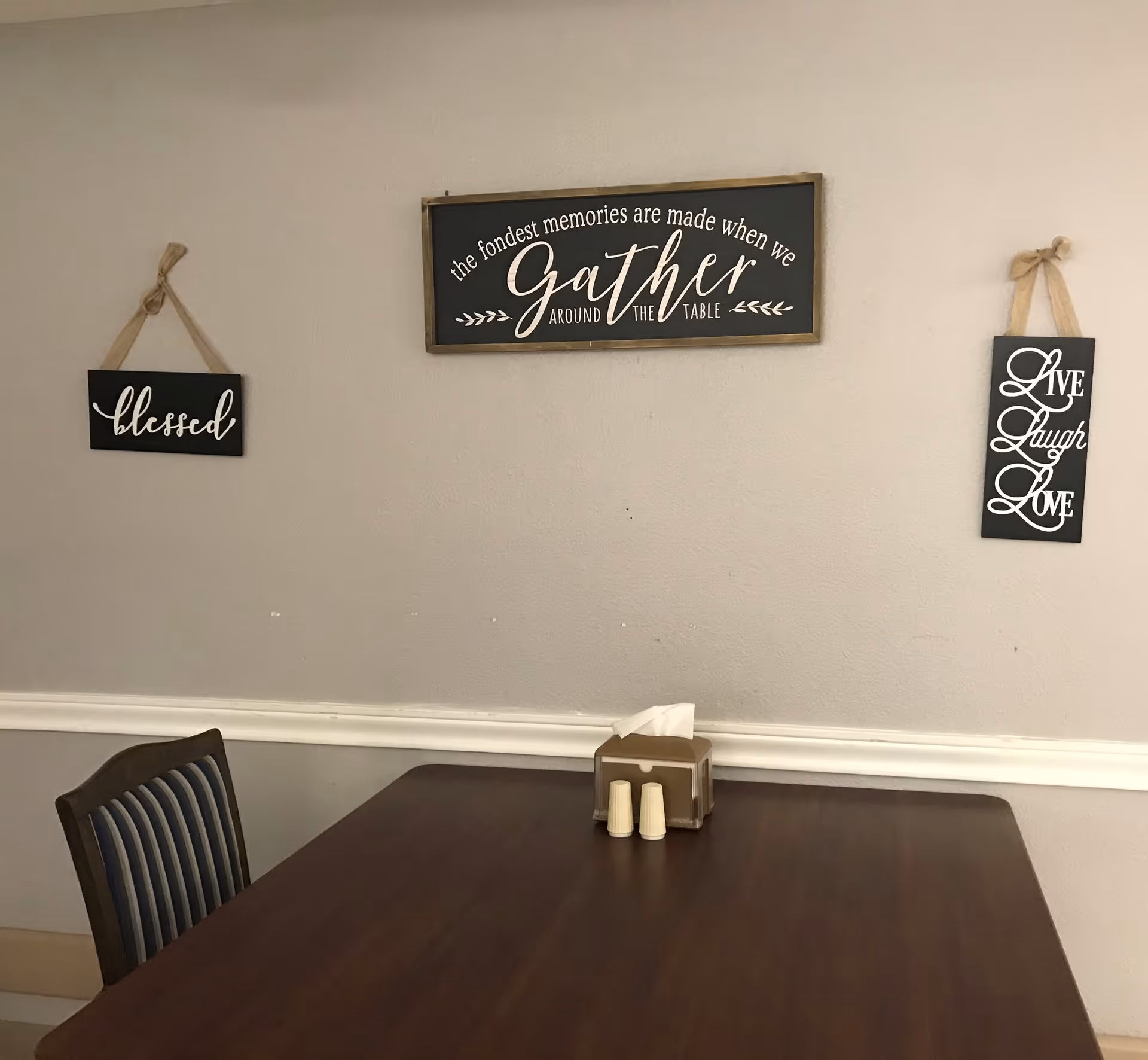 A wooden dining table with a striped chair is positioned against a beige wall. On the wall, there are three decorative signs with inspirational quotes: 'blessed', 'the fondest memories are made when we gather around the table', and 'Live Laugh Love'. On the table, there is a tissue box and two small containers.