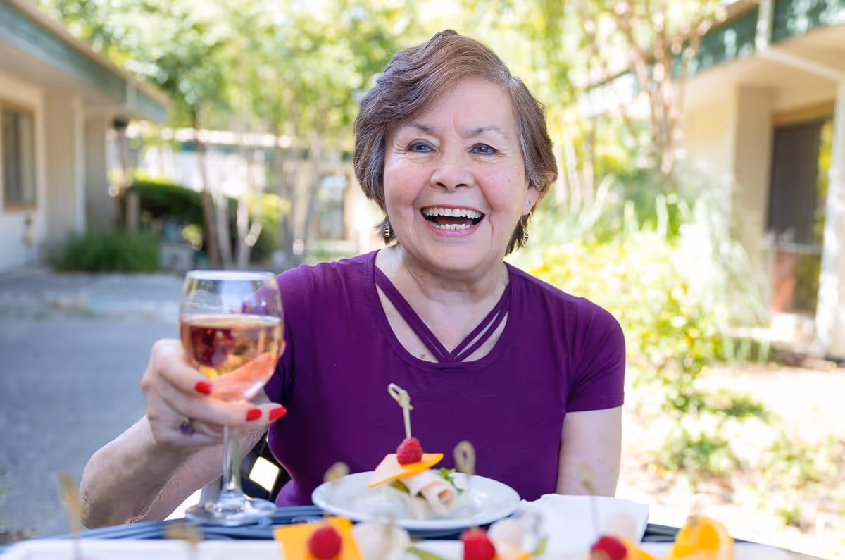 A smiling elderly woman wearing a purple top is sitting outdoors at a table with appetizers and holding up a glass of rosé wine in a toast. The background shows greenery and parts of a building.