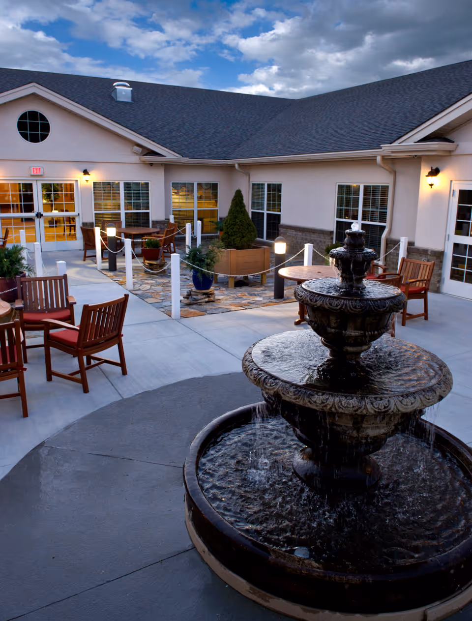 Outdoor patio area at a senior living facility with a multi-tiered water fountain in the foreground, wooden chairs with red cushions, round tables, potted plants, and a building with large windows and doors in the background under a cloudy sky.