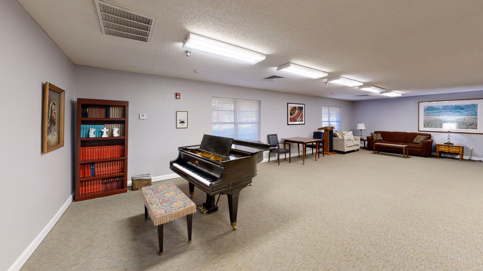 A spacious common room with a black grand piano and a cushioned piano bench in the center. To the left, there is a bookshelf filled with books and decorative items, and a framed picture of a dog on the wall. The room has beige carpet, light gray walls, and several windows letting in natural light. At the far end, there are two sofas, a wooden coffee table, a side table with a lamp, and framed artwork on the walls. The ceiling has multiple fluorescent light fixtures.