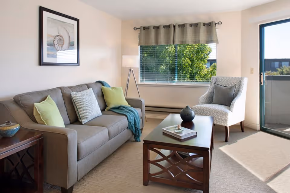 A bright living room with a beige sofa adorned with green and striped pillows, a patterned armchair with a gray pillow, a wooden coffee table with decorative items and a book, a floor lamp, a framed seashell artwork on the wall, and a large window with a view of green trees. There is also a sliding glass door leading to a balcony.
