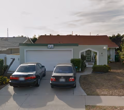Single-story residential house with a red roof and green exterior walls. Two cars are parked in the driveway in front of a double garage door. There is a small front yard with some bushes and a walkway leading to the front door.