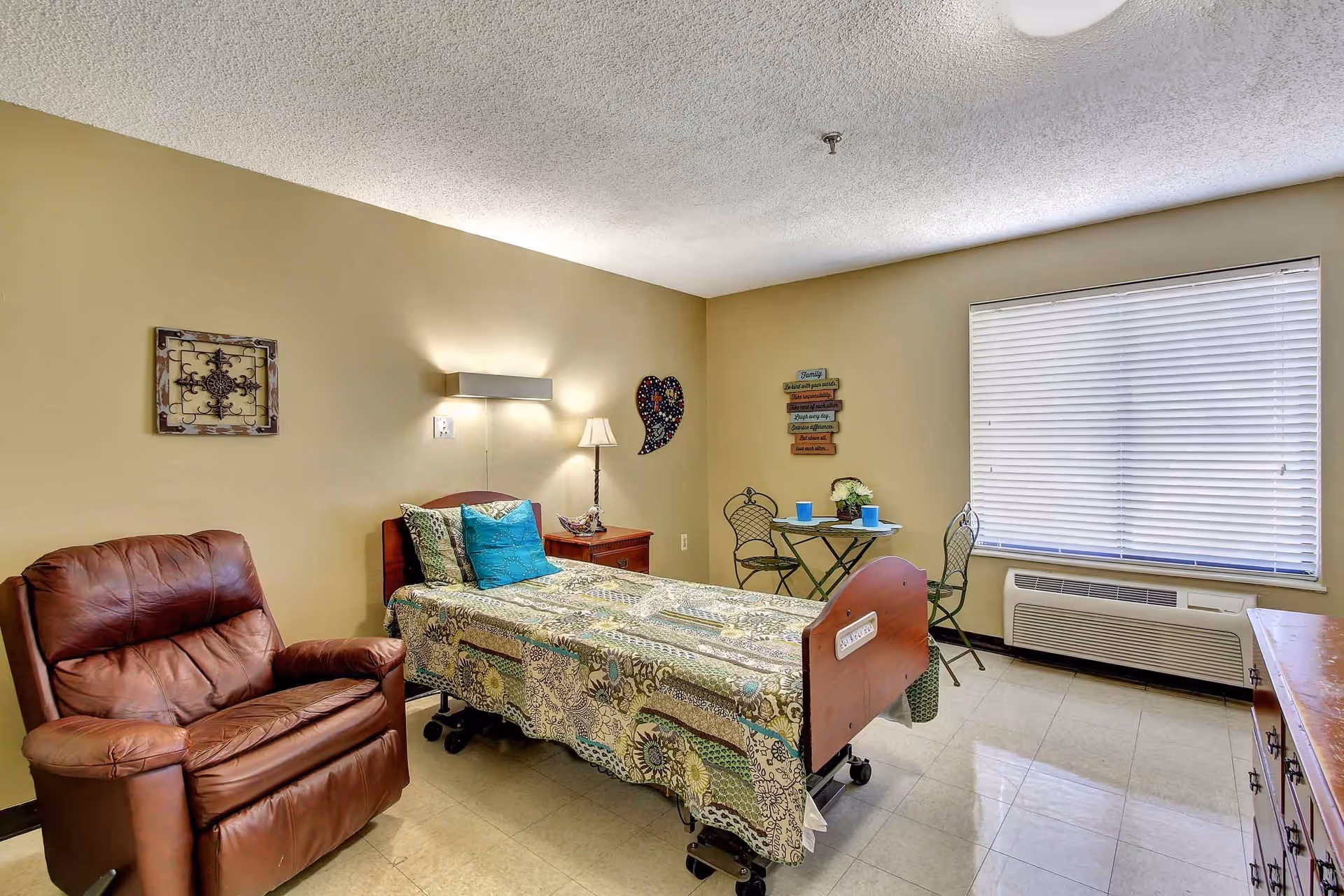 Assisted-living bedroom with a single bed, bedside table and lamp, a leather recliner, small bistro table with two chairs, and a window with blinds.