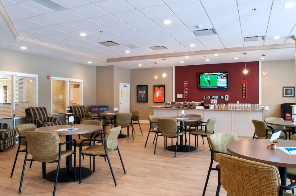 A spacious dining area in a senior living facility with round tables and chairs arranged neatly. The room features a bar counter with glasses and a television mounted on a red accent wall showing a sports program. Comfortable armchairs are placed near the windows on the left side, and the ceiling has recessed lighting.