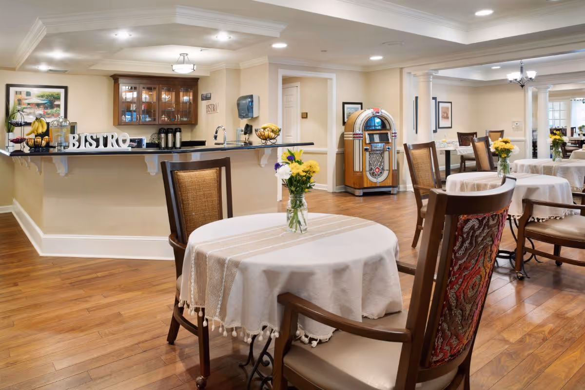A cozy dining area in a senior living facility with round tables covered in white tablecloths and small flower vases. Wooden chairs surround the tables. In the background, there is a counter with a 'BISTRO' sign, coffee dispensers, and a fruit basket. A vintage jukebox is positioned against the wall, and the room features warm lighting and wooden flooring.