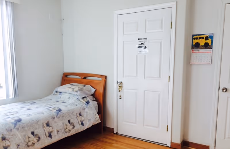 A simple bedroom with a single wooden bed covered with a floral patterned bedspread and pillow. There is a white door with a safety notice on it and a wall calendar featuring a yellow school bus. The room has light-colored walls and a window with vertical blinds letting in natural light.