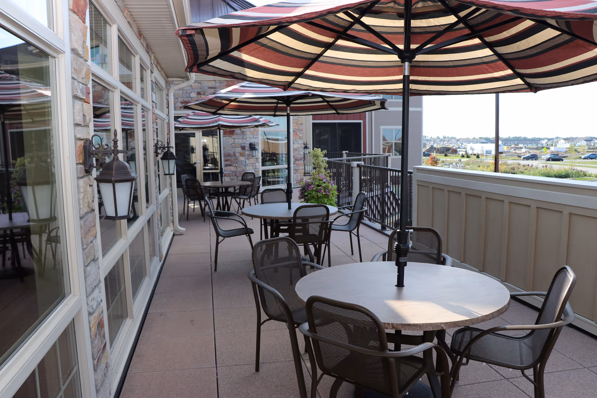Outdoor patio area with round tables and metal chairs under large striped umbrellas. The patio is adjacent to a building with stone and siding exterior walls and large windows. There are decorative wall-mounted lanterns along the building. In the background, a parking lot and distant buildings are visible.