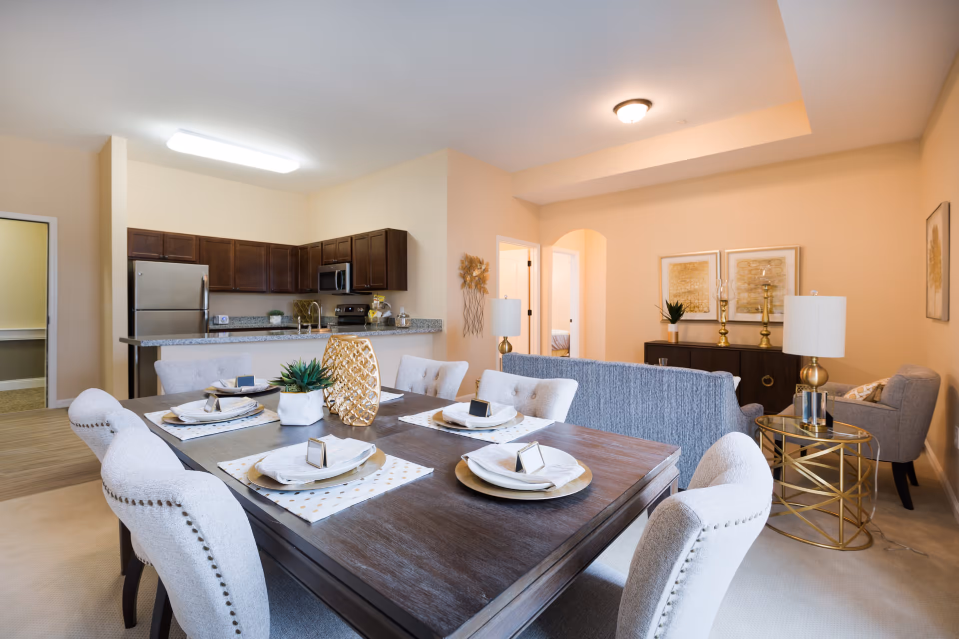 A bright and modern open-concept living space featuring a dining area with a wooden table set for four, upholstered chairs, and decorative centerpieces. In the background, there is a kitchen with dark wood cabinets, stainless steel appliances including a refrigerator and microwave, and a granite countertop. Adjacent to the dining area is a living room with a gray sofa, armchair, gold-accented side table with a lamp, and framed artwork on the walls. The space has beige walls and carpeted flooring.