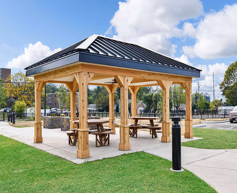 Outdoor wooden pavilion with a black metal roof, containing picnic tables and benches, situated on a concrete pad surrounded by green grass and a parking area in the background under a partly cloudy sky.