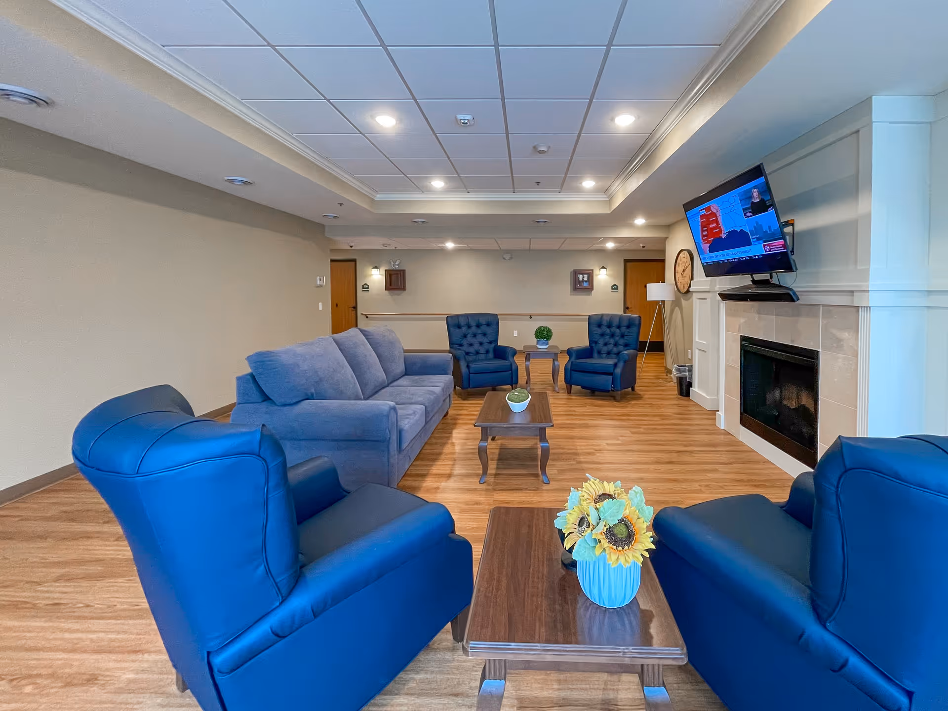 A cozy living room area in a senior living facility with blue armchairs and a blue sofa arranged around wooden coffee tables. A flat-screen TV is mounted above a fireplace on the right wall. The room has wood flooring, recessed ceiling lights, and neutral-colored walls.