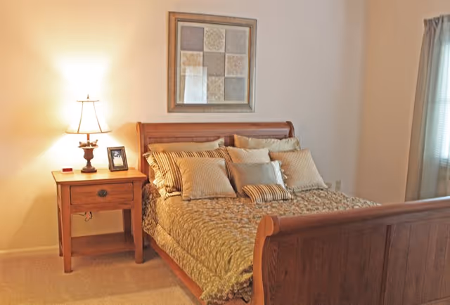 Cozy bedroom with a wooden sleigh bed dressed in patterned bedding and multiple pillows, a nightstand with a lamp and framed photo, and wall art above the bed.