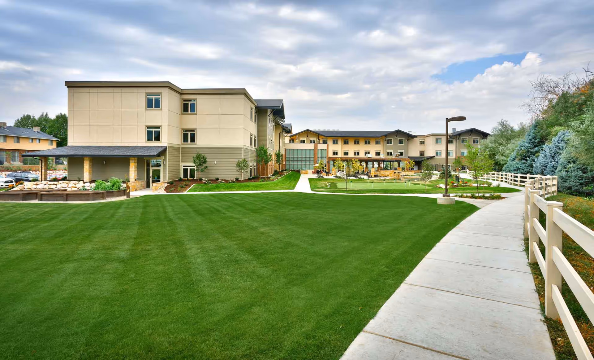 Front exterior of a three-story assisted living building with a wide manicured lawn and a curved sidewalk leading toward the entrance.