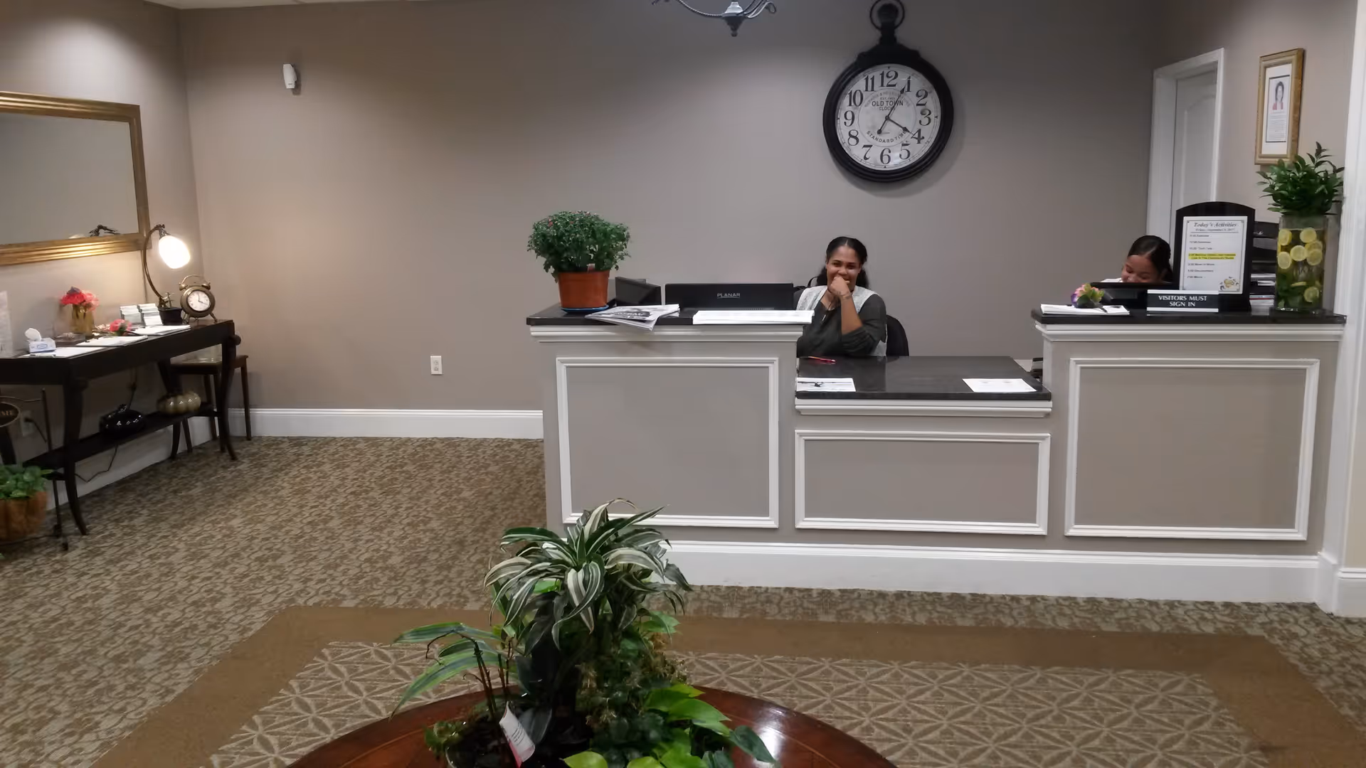 Reception area of Susan S Bailis Assisted Living with two receptionists behind a white counter. There is a large round clock on the wall behind them, a table with plants in the foreground, and a side table with a lamp, clock, and flowers on the left side.