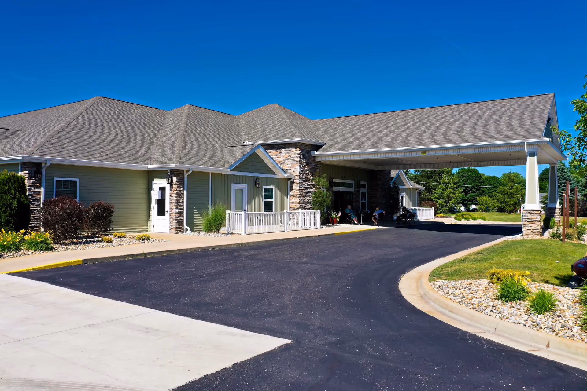 Exterior view of a single-story assisted living facility building with a covered entrance driveway, stone and siding facade, landscaped surroundings, and a clear blue sky.