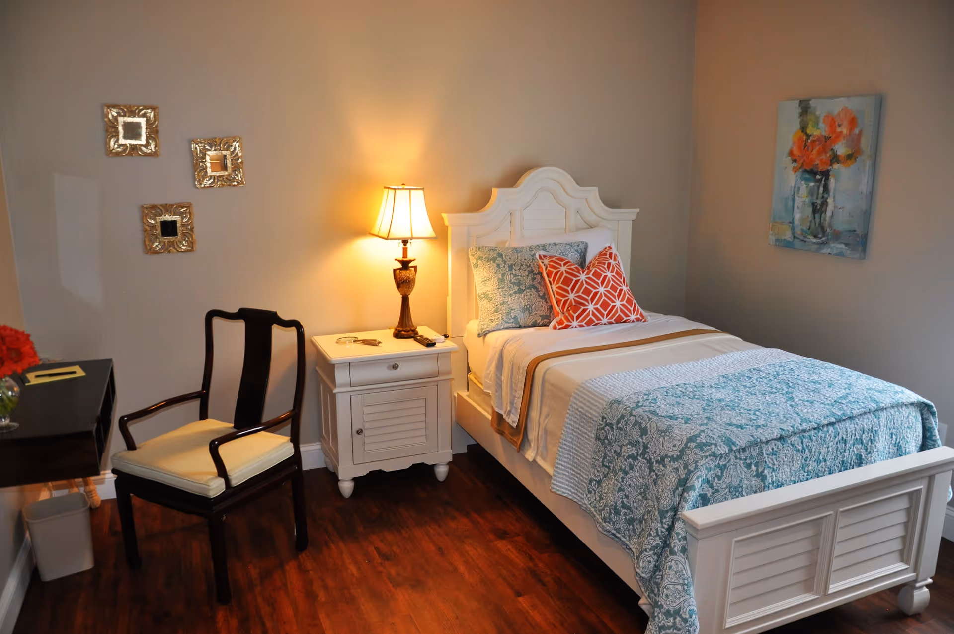 A cozy bedroom with a white wooden bed featuring blue and orange patterned pillows and a blue floral bedspread. Next to the bed is a white nightstand with a lamp emitting warm light. A dark wooden chair with a cream cushion is positioned near a small black desk with a vase of red flowers. The walls are decorated with three small ornate mirrors and a painting of orange flowers in a vase. The floor is wooden.