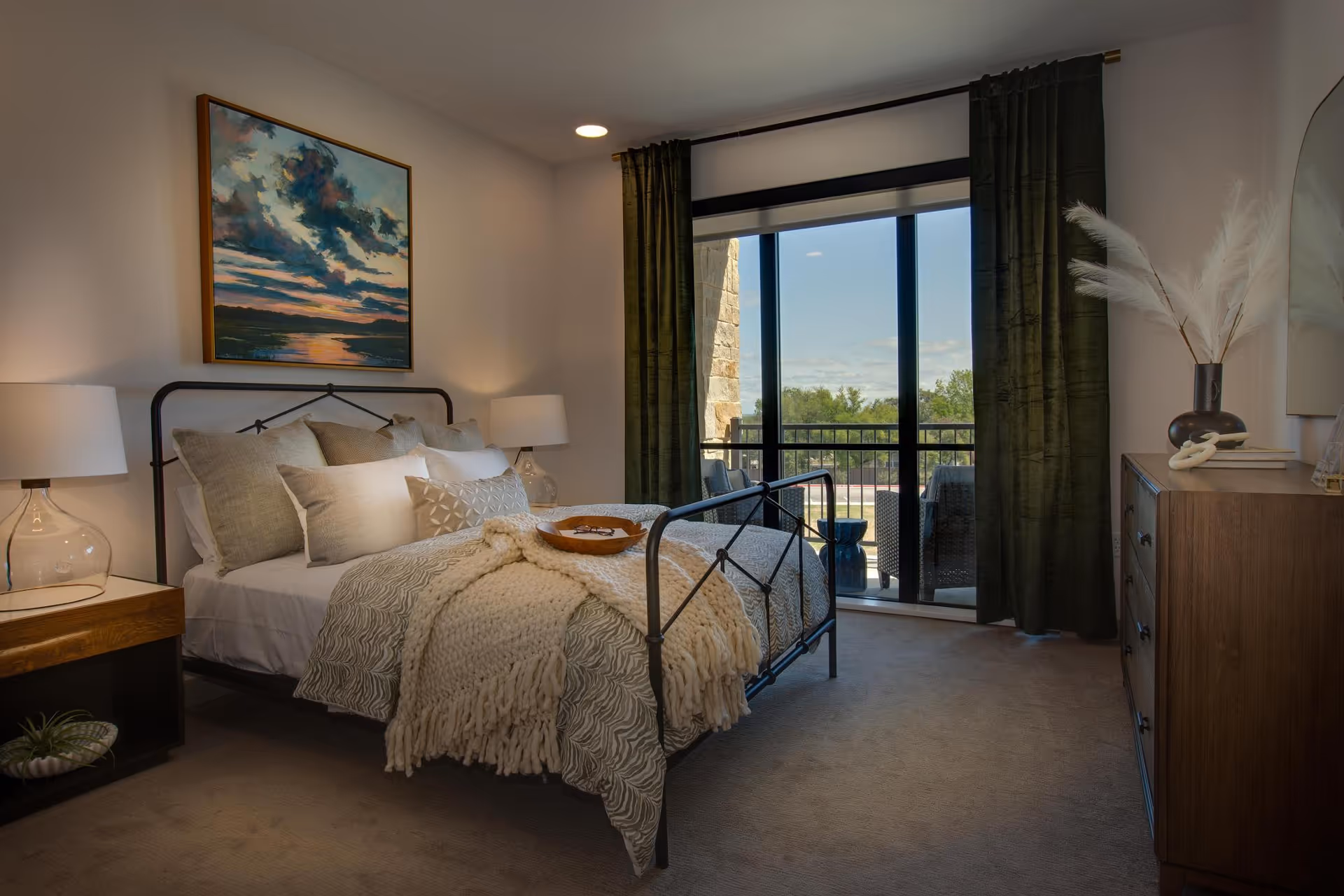 A cozy bedroom with a metal frame bed adorned with multiple pillows and a textured blanket. Two bedside tables with glass lamps flank the bed. A large painting of a sunset over water hangs above the bed. Floor-to-ceiling windows with dark curtains open to a balcony with outdoor seating and a view of trees and a partly cloudy sky. A wooden dresser with decorative items is on the right side of the room.