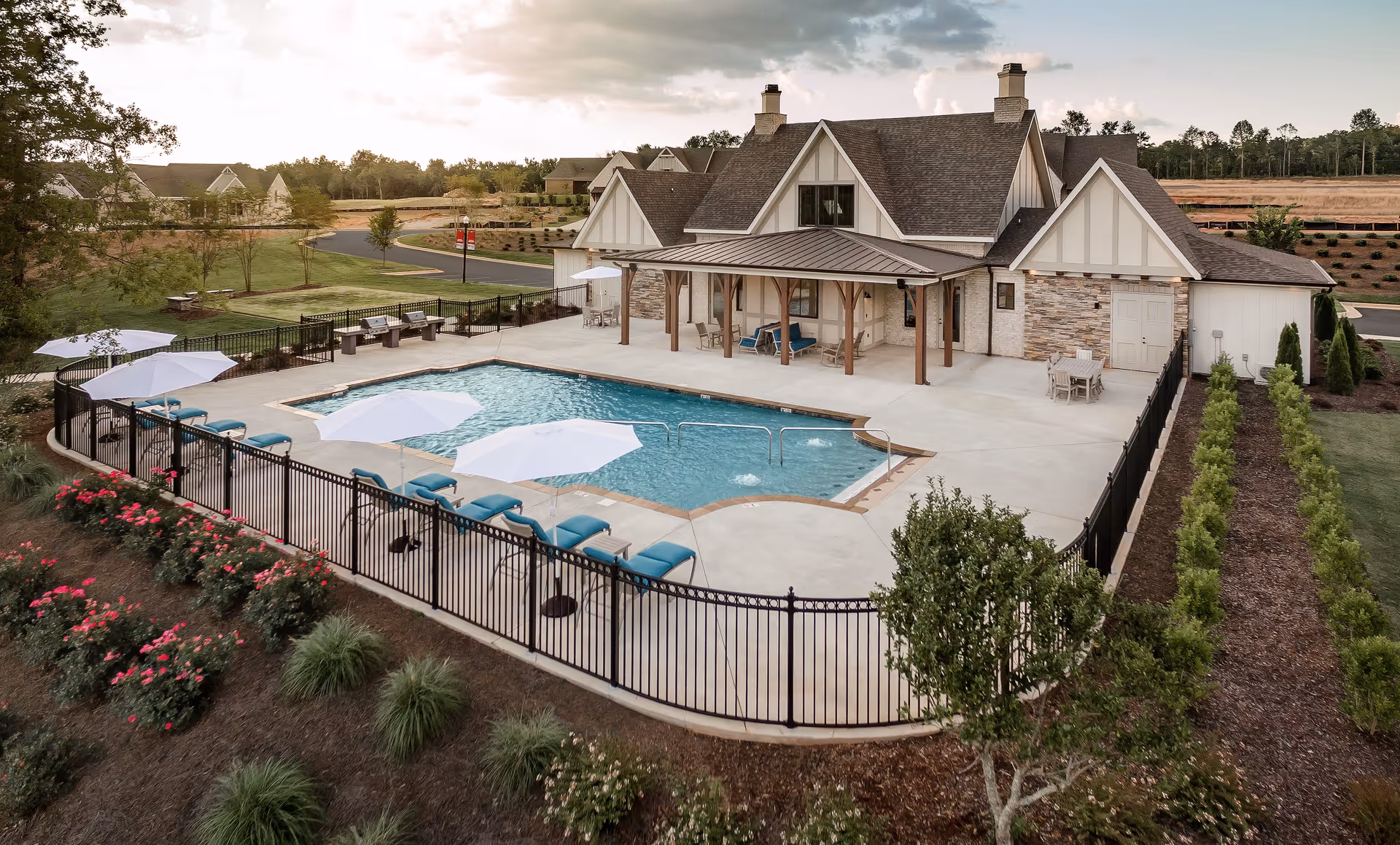 Outdoor swimming pool area surrounded by a black metal fence with several lounge chairs and white umbrellas. Behind the pool is a large building with a covered patio, stone and wood exterior, and a sloped roof. The surrounding area includes landscaped gardens with bushes and flowers, a paved road, and trees in the background under a cloudy sky.
