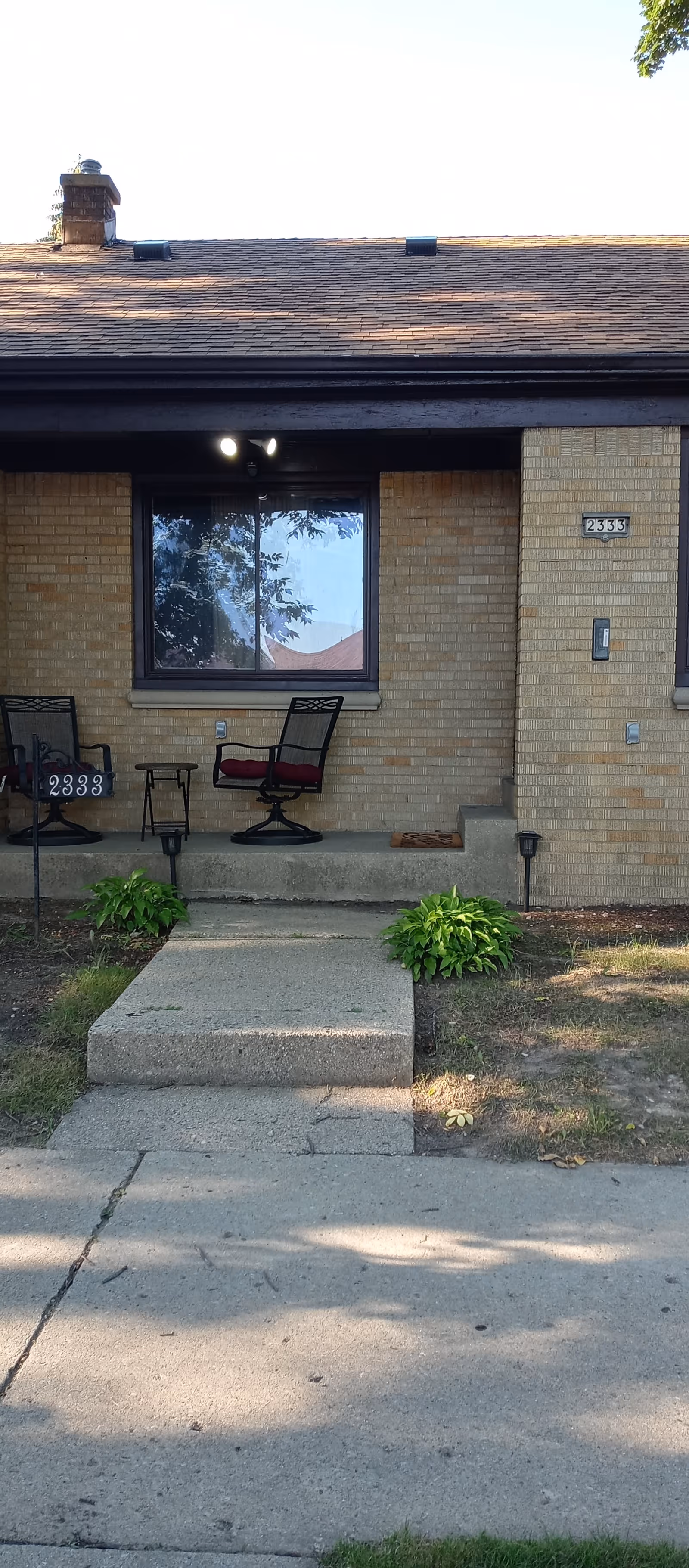 Front view of a single-story brick building with a small concrete porch. Two black metal chairs with red cushions and a small table are on the porch. The building has a window with reflections of trees and a house. The address number 2333 is visible on the wall and on a small sign near the porch. There are small green plants on either side of the concrete walkway leading to the porch.