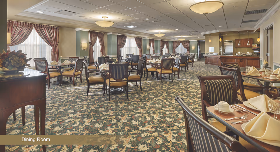 A spacious dining room with multiple tables set for a meal, featuring folded napkins, cups, and silverware. The room has patterned carpet, large windows with red curtains, and warm lighting fixtures on the ceiling and walls. There is a serving area with a counter and kitchen appliances in the background.
