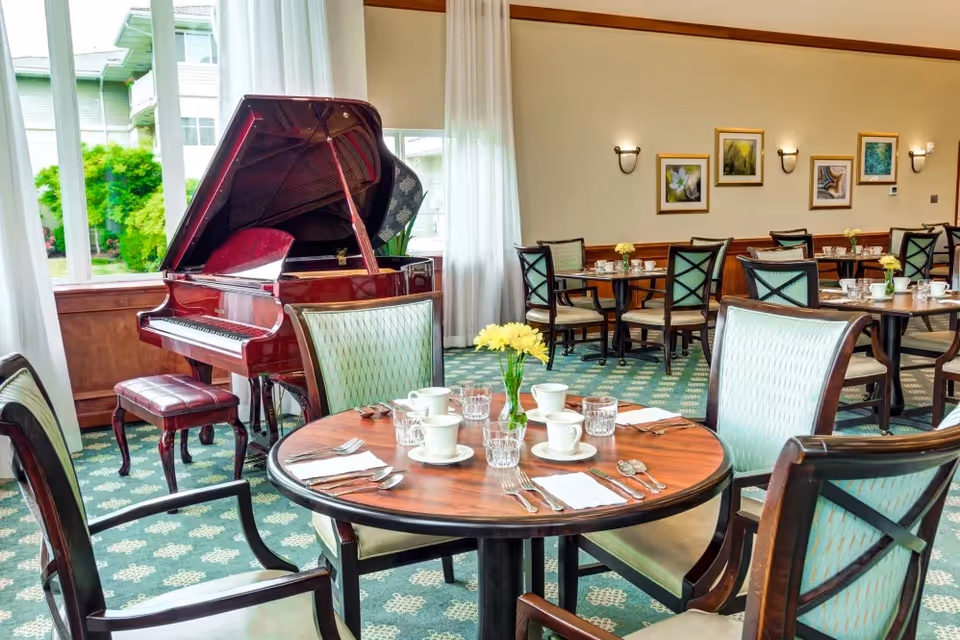 A bright dining room with round wooden tables set with white cups, glasses, and silverware. The room features green patterned carpet, light green cushioned chairs with wooden frames, and large windows with white curtains letting in natural light. A polished red grand piano with a matching bench is positioned near the windows. The walls are decorated with framed artwork and wall sconces.