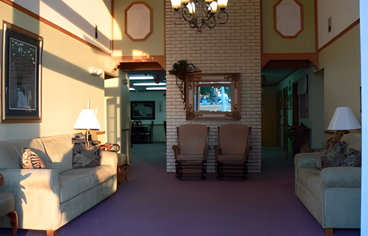 A cozy living room area with two beige sofas facing each other, each with decorative pillows and lamps on side tables. In the center, there are two wooden rocking chairs placed in front of a white brick wall with a mirror and a small plant. The room is softly lit by natural light coming from the left side, casting shadows on the walls.